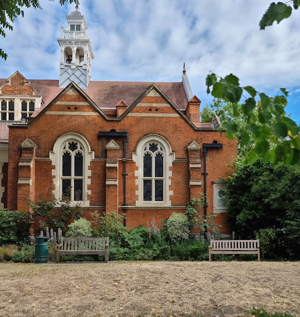 A tranquil small garden scene showing a narrow, paved walkway leading towards a church at the distance, framed by leafless trees and green shrubbery. The garden features neatly maintained grass lawns 